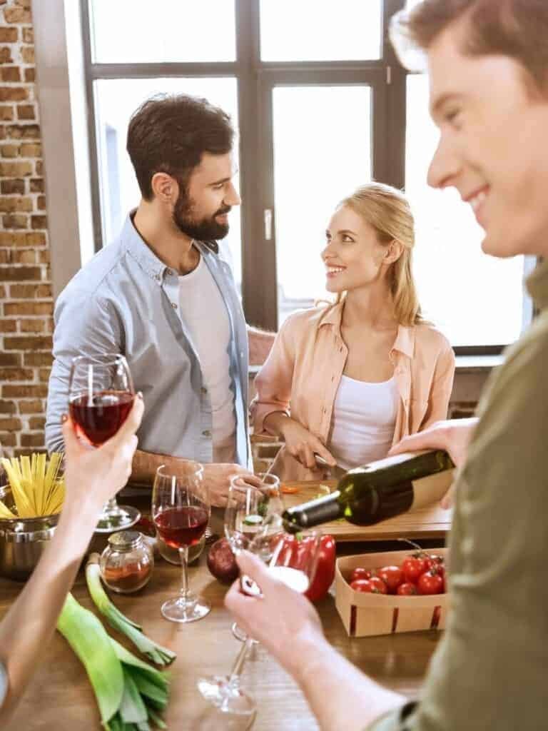 A smiling man and woman in a kitchen with a party taking place. They are both dressed casually and in the foreground there is a hand holding a glass of wine as a man pours his own wine from a bottle. Party food is present on the table.